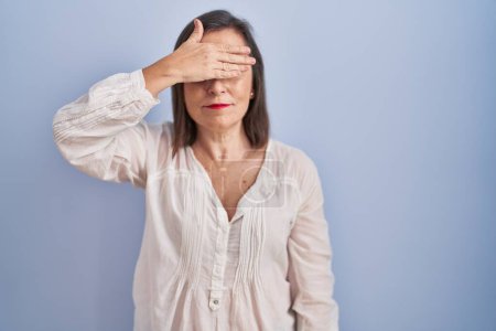 Foto de Middle age hispanic woman standing over blue background covering eyes with hand, looking serious and sad. sightless, hiding and rejection concept - Imagen libre de derechos