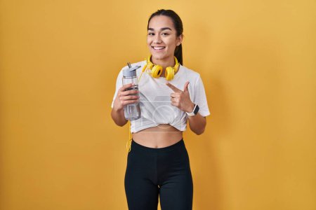 Foto de Young south asian woman wearing sportswear drinking water cheerful with a smile on face pointing with hand and finger up to the side with happy and natural expression - Imagen libre de derechos