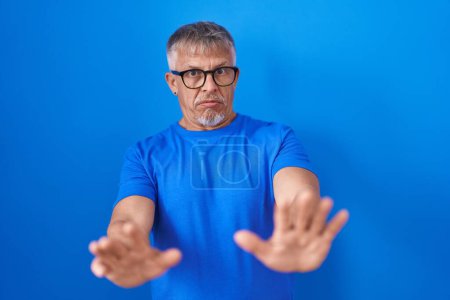 Photo for Hispanic man with grey hair standing over blue background moving away hands palms showing refusal and denial with afraid and disgusting expression. stop and forbidden. - Royalty Free Image