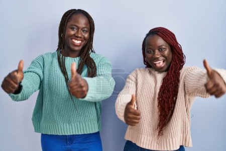 Téléchargez les photos : Deux femmes africaines debout sur fond bleu approuvant de faire un geste positif avec la main, le pouce levé souriant et heureux pour le succès. geste gagnant. - en image libre de droit