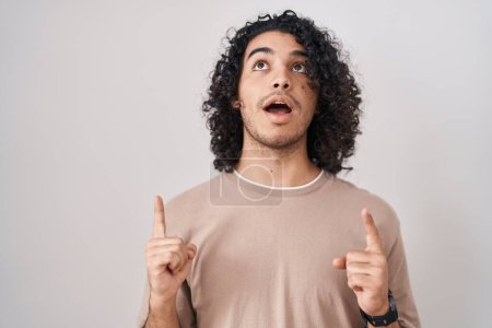 Foto de Hispanic man with curly hair standing over white background amazed and surprised looking up and pointing with fingers and raised arms. - Imagen libre de derechos