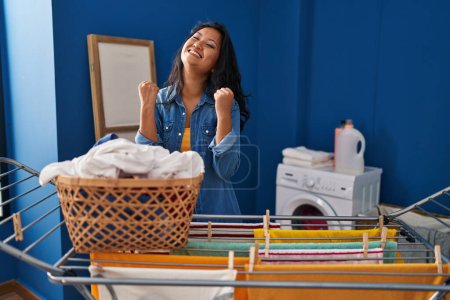 Foto de Young asian woman hanging clothes at clothesline very happy and excited doing winner gesture with arms raised, smiling and screaming for success. celebration concept. - Imagen libre de derechos