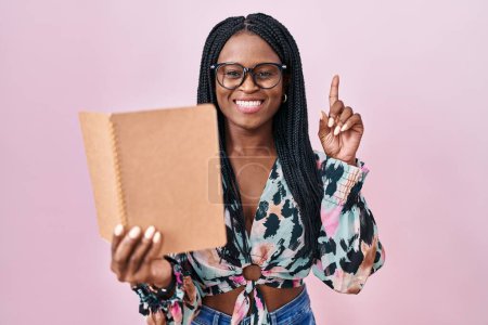 Foto de African woman with braids reading a book surprised with an idea or question pointing finger with happy face, number one - Imagen libre de derechos