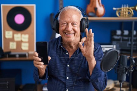 Foto de Senior man with grey hair showing smartphone screen at music studio doing ok sign with fingers, smiling friendly gesturing excellent symbol - Imagen libre de derechos