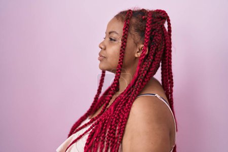 Foto de Mujer afroamericana con cabello trenzado de pie sobre fondo rosa mirando a un lado, pose de perfil relajado con rostro natural y sonrisa confiada. - Imagen libre de derechos
