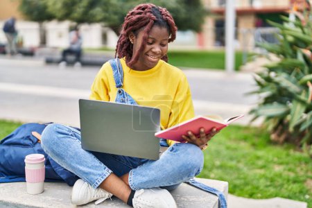 Foto de African american woman student using laptop reading book at campus park - Imagen libre de derechos