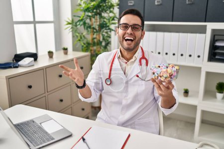 Photo for Young hispanic doctor man with beard holding candy celebrating victory with happy smile and winner expression with raised hands - Royalty Free Image