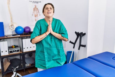 Photo for Young hispanic woman wearing physiotherapist uniform standing at clinic begging and praying with hands together with hope expression on face very emotional and worried. begging. - Royalty Free Image