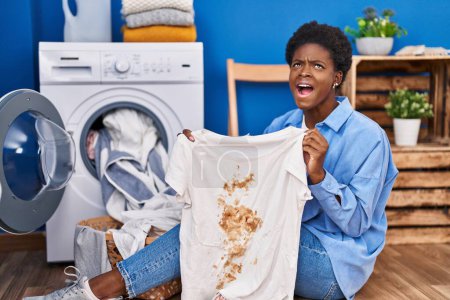 Photo for African american woman holding t shirt with dirty stain angry and mad screaming frustrated and furious, shouting with anger looking up. - Royalty Free Image