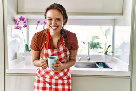 Téléchargez les photos : Young beautiful hispanic woman wearing apron drinking coffee at the kitchen - en image libre de droit