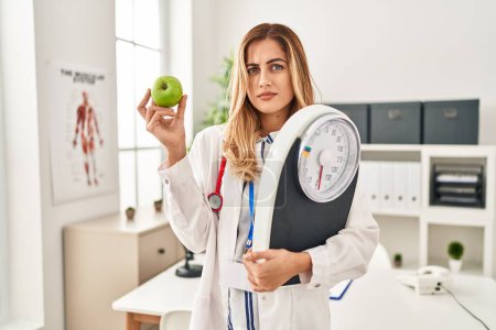Foto de Joven doctora rubia sosteniendo máquina de pesaje y manzana verde expresión despistada y confusa. concepto de duda. - Imagen libre de derechos