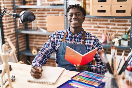 Téléchargez les photos : African american man artist drawing on notebook reading book at art studio - en image libre de droit