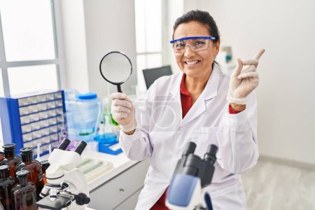 Téléchargez les photos : Moyen Age femme hispanique travaillant au laboratoire scientifique tenant loupe sourire heureux pointant avec la main et le doigt vers le côté - en image libre de droit