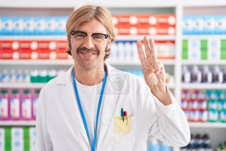 Photo for Caucasian man with mustache working at pharmacy drugstore showing and pointing up with fingers number three while smiling confident and happy. - Royalty Free Image