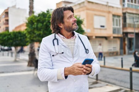 Téléchargez les photos : Moyen âge homme médecin souriant confiant en utilisant smartphone à la rue - en image libre de droit