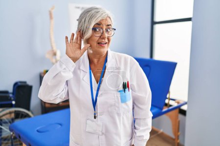 Photo for Middle age woman with grey hair working at pain recovery clinic smiling with hand over ear listening an hearing to rumor or gossip. deafness concept. - Royalty Free Image