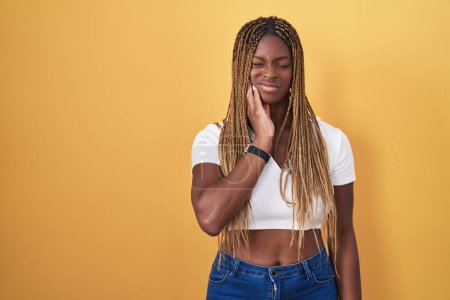Téléchargez les photos : African american woman with braided hair standing over yellow background touching mouth with hand with painful expression because of toothache or dental illness on teeth. dentist - en image libre de droit