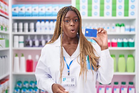 Photo for African american woman with braided hair working at pharmacy drugstore holding credit card scared and amazed with open mouth for surprise, disbelief face - Royalty Free Image