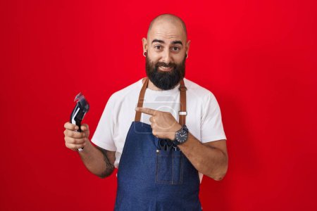 Foto de Young hispanic man with beard and tattoos wearing barber apron holding razor pointing to the back behind with hand and thumbs up, smiling confident - Imagen libre de derechos