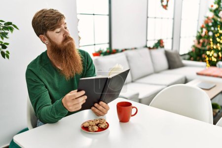 Téléchargez les photos : Jeune rousse homme prendre le petit déjeuner assis près de l'arbre de Noël à la maison - en image libre de droit