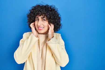 Photo for Young brunette woman with curly hair standing over blue background covering ears with fingers with annoyed expression for the noise of loud music. deaf concept. - Royalty Free Image