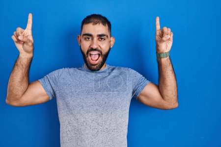 Foto de Hombre de Oriente Medio con barba de pie sobre fondo azul sonriendo sorprendido y sorprendido y apuntando hacia arriba con los dedos y los brazos levantados. - Imagen libre de derechos