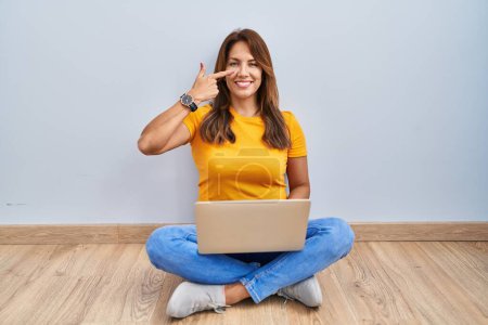 Téléchargez les photos : Hispanic woman using laptop sitting on the floor at home pointing with hand finger to face and nose, smiling cheerful. beauty concept - en image libre de droit