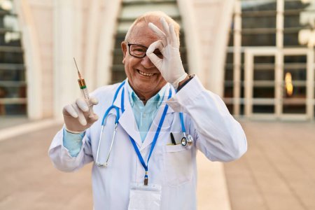 Foto de Senior doctor with grey hair holding syringe smiling happy doing ok sign with hand on eye looking through fingers - Imagen libre de derechos