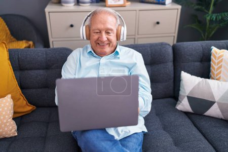 Photo for Middle age grey-haired man using laptop and headphones sitting on sofa at home - Royalty Free Image