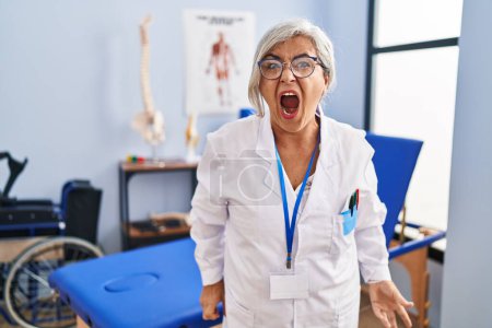 Photo for Middle age woman with grey hair working at pain recovery clinic angry and mad screaming frustrated and furious, shouting with anger. rage and aggressive concept. - Royalty Free Image
