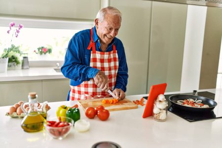 Foto de Hombre mayor sonriendo confiado cocinar y ver receta en línea en la cocina - Imagen libre de derechos