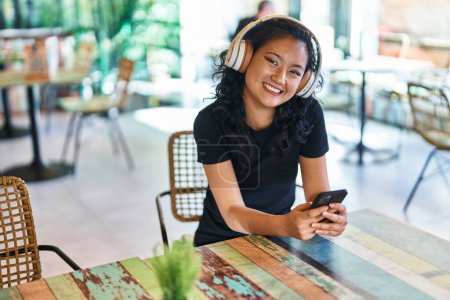 Téléchargez les photos : Young chinese woman using smartphone and headphones sitting on table at restaurant - en image libre de droit