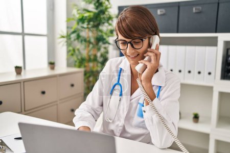 Téléchargez les photos : Jeune belle hispanique femme médecin en utilisant ordinateur portable parler au téléphone à la clinique - en image libre de droit