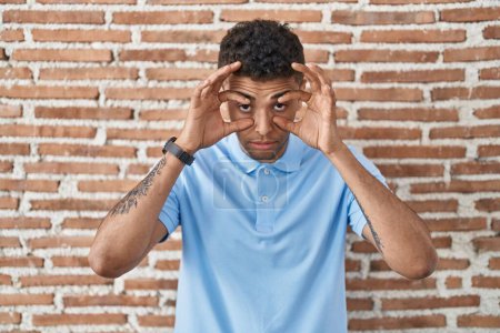 Téléchargez les photos : Brazilian young man standing over brick wall trying to open eyes with fingers, sleepy and tired for morning fatigue - en image libre de droit