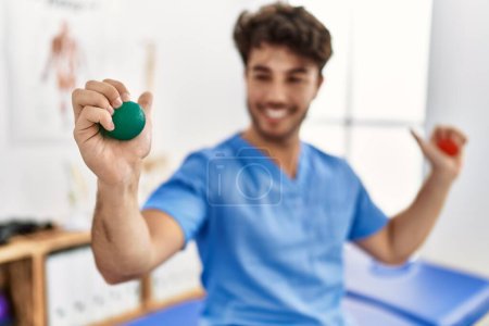 Photo for Young hispanic man wearing physio therapist uniform using anti stress ball at clinic - Royalty Free Image