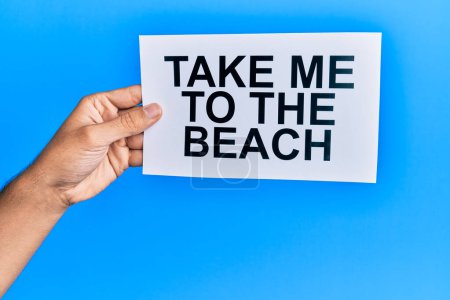 Foto de Hand of caucasian man holding paper with take me to the beach message over isolated blue background - Imagen libre de derechos