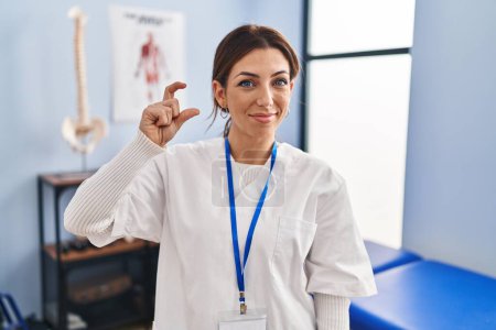 Photo for Young brunette woman working at pain recovery clinic smiling and confident gesturing with hand doing small size sign with fingers looking and the camera. measure concept. - Royalty Free Image