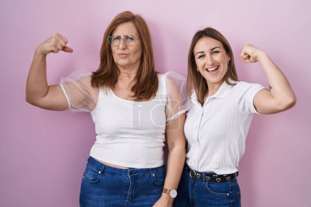 Foto de Madre e hija hispanas vistiendo camiseta blanca casual sobre fondo rosa persona fuerte mostrando músculo del brazo, confiado y orgulloso de poder - Imagen libre de derechos