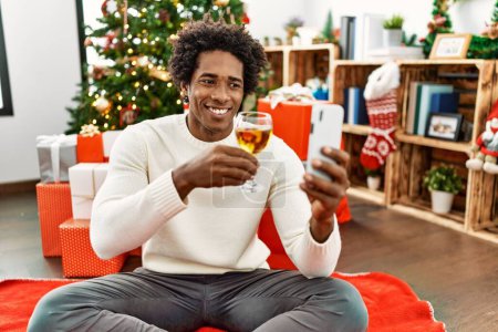 Foto de Joven afroamericano hombre hacer selfie por el teléfono inteligente sentado en el suelo por árbol de Navidad en casa. - Imagen libre de derechos
