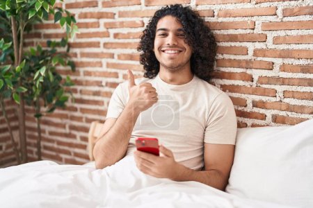 Foto de Hispanic man with curly hair using smartphone sitting on the bed smiling happy and positive, thumb up doing excellent and approval sign - Imagen libre de derechos
