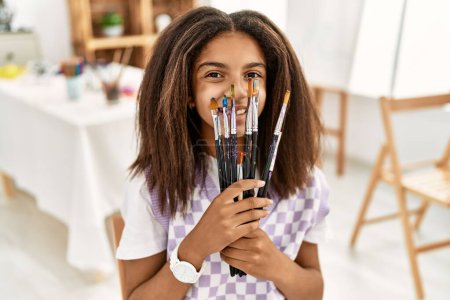 Photo for African american girl smiling confident covering mouth with paintbrushes at art school - Royalty Free Image