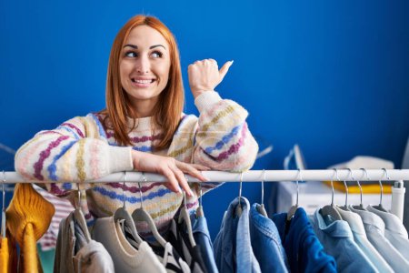 Photo for Young woman searching clothes on clothing rack pointing thumb up to the side smiling happy with open mouth - Royalty Free Image