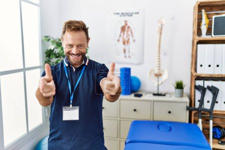 Foto de Middle age physiotherapist man working at pain recovery clinic pointing fingers to camera with happy and funny face. good energy and vibes. - Imagen libre de derechos