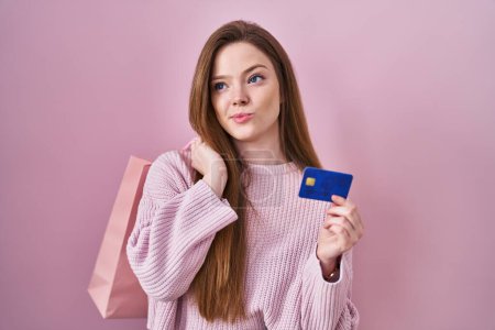 Foto de Joven mujer caucásica sosteniendo bolsa de compras y tarjeta de crédito sonriendo mirando a un lado y mirando hacia otro pensando. - Imagen libre de derechos