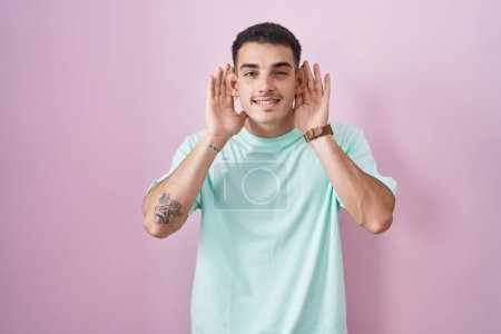 Photo for Handsome hispanic man standing over pink background trying to hear both hands on ear gesture, curious for gossip. hearing problem, deaf - Royalty Free Image