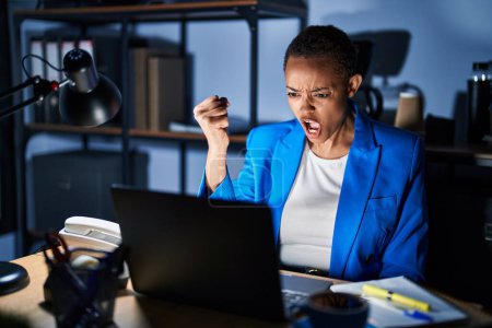 Téléchargez les photos : Beautiful african american woman working at the office at night angry and mad raising fist frustrated and furious while shouting with anger. rage and aggressive concept. - en image libre de droit