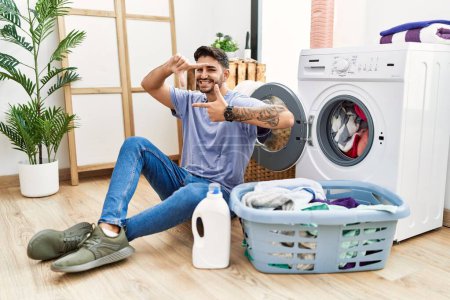 Foto de Young hispanic man putting dirty laundry into washing machine smiling making frame with hands and fingers with happy face. creativity and photography concept. - Imagen libre de derechos