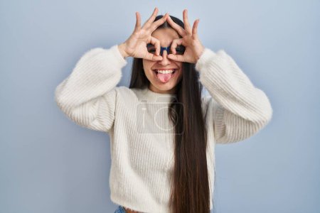 Foto de Young hispanic woman wearing casual sweater over blue background doing ok gesture like binoculars sticking tongue out, eyes looking through fingers. crazy expression. - Imagen libre de derechos