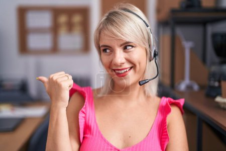 Photo for Young caucasian woman wearing call center agent headset smiling with happy face looking and pointing to the side with thumb up. - Royalty Free Image