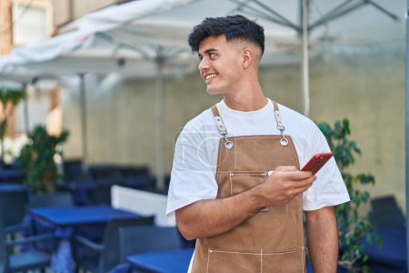 Téléchargez les photos : Jeune homme hispanique serveur souriant confiant en utilisant un smartphone à la terrasse du café - en image libre de droit